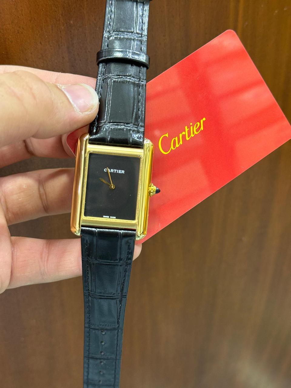 A Cartier Tank watch with a gold-tone case, black dial, and black alligator strap, held by a hand next to its signature red Cartier box.